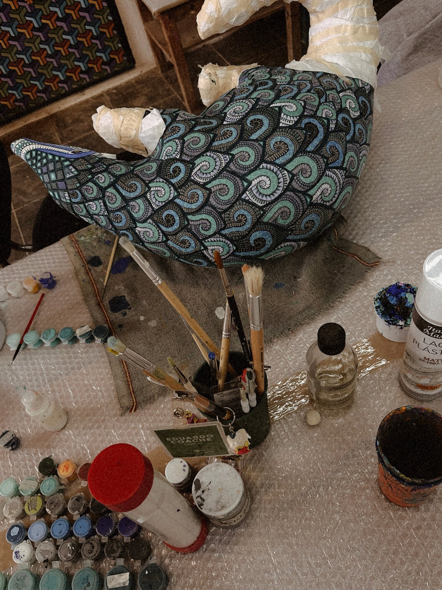 Jacobo and María Ángeles, Mexico — Artisan studio workspace with ornate patterned chair, paint palettes, brushes, and craft materials on concrete floor.