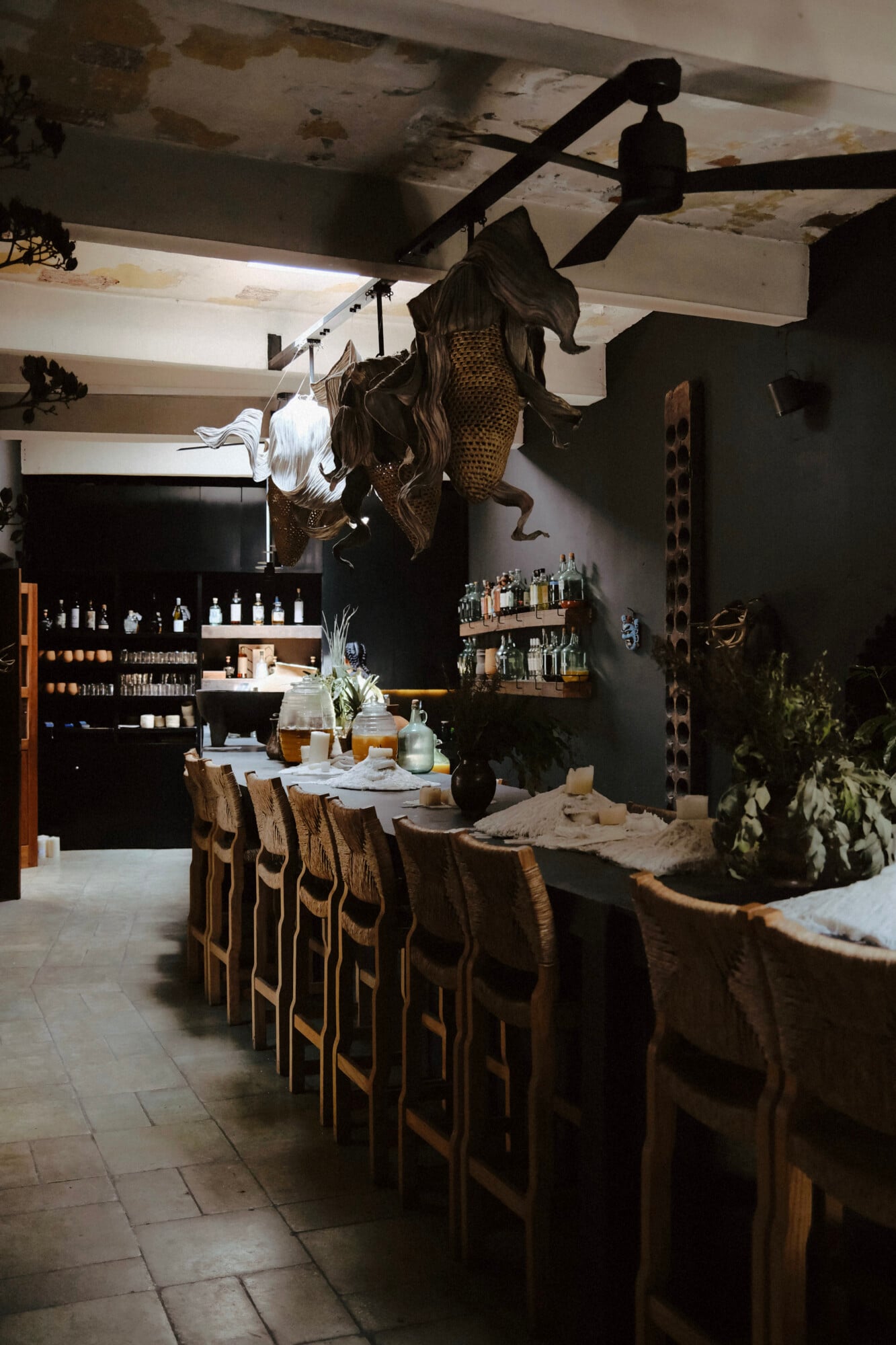 Criollo Rstaurante — Criollo Restaurant, Mexico — Dark dining room with sculptural clay figures, woven chairs, exposed beams, brass pendant lighting, and botanical arrangements.