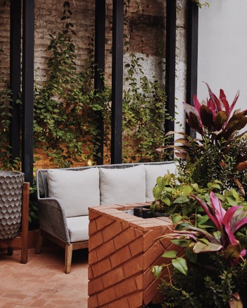 Campos Polanco, Mexico — Brick courtyard with woven lounge chair, white cushioned seating, climbing vines on black trellis, and vibrant tropical plants in afternoon light.