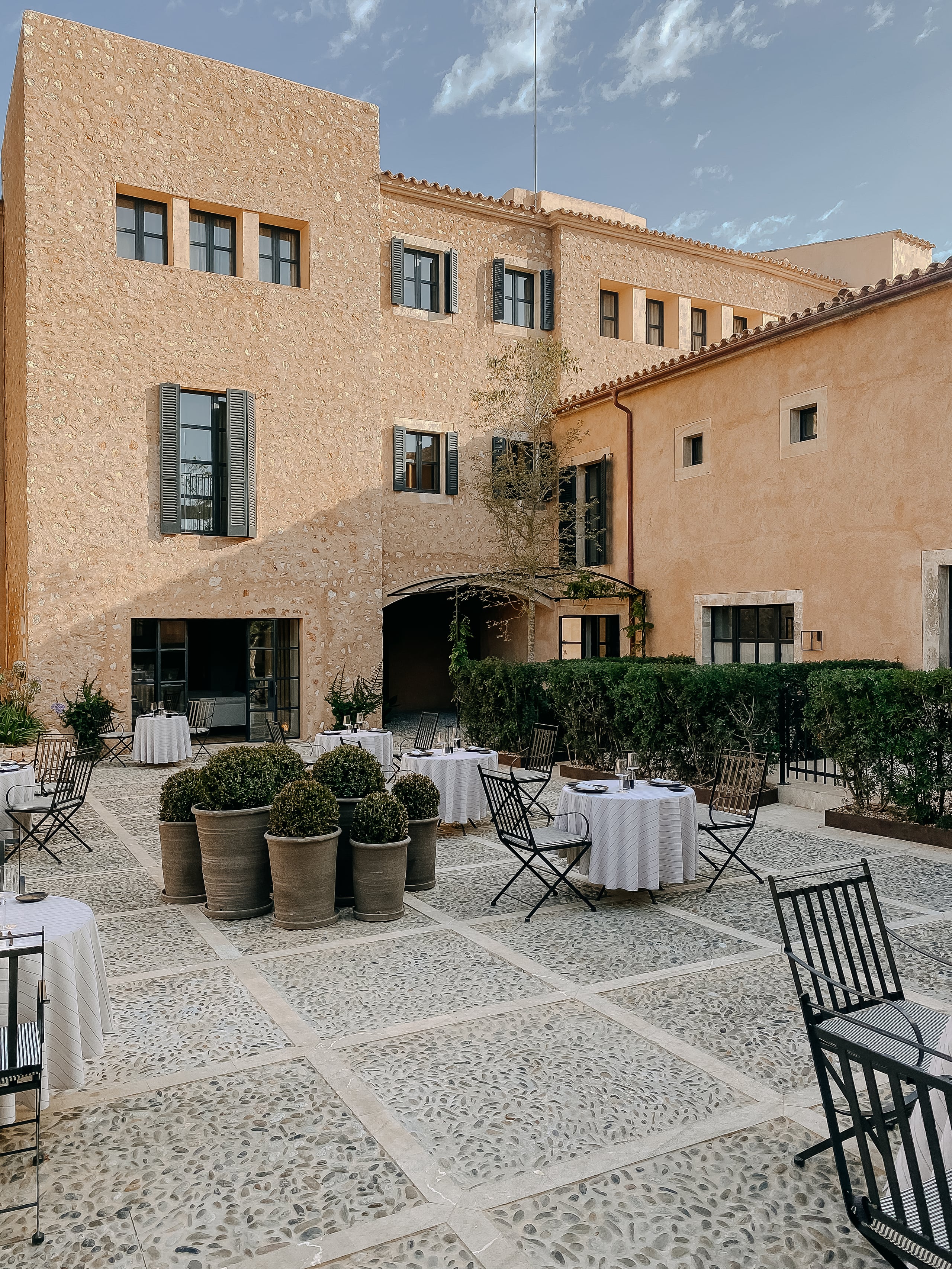 Courtyard dining at Can Ferrereta with terracotta buildings, geometric stone flooring, potted topiaries, and black chairs beneath Mediterranean light.