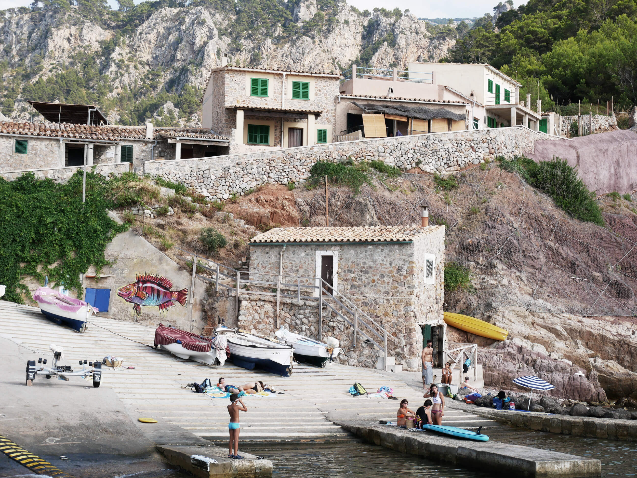 Beaches In Mallorca — Mediterranean beach cove with stone boathouse, moored fishing boats, limestone cliffs, terraced villas with green shutters, swimmers in shallow water.