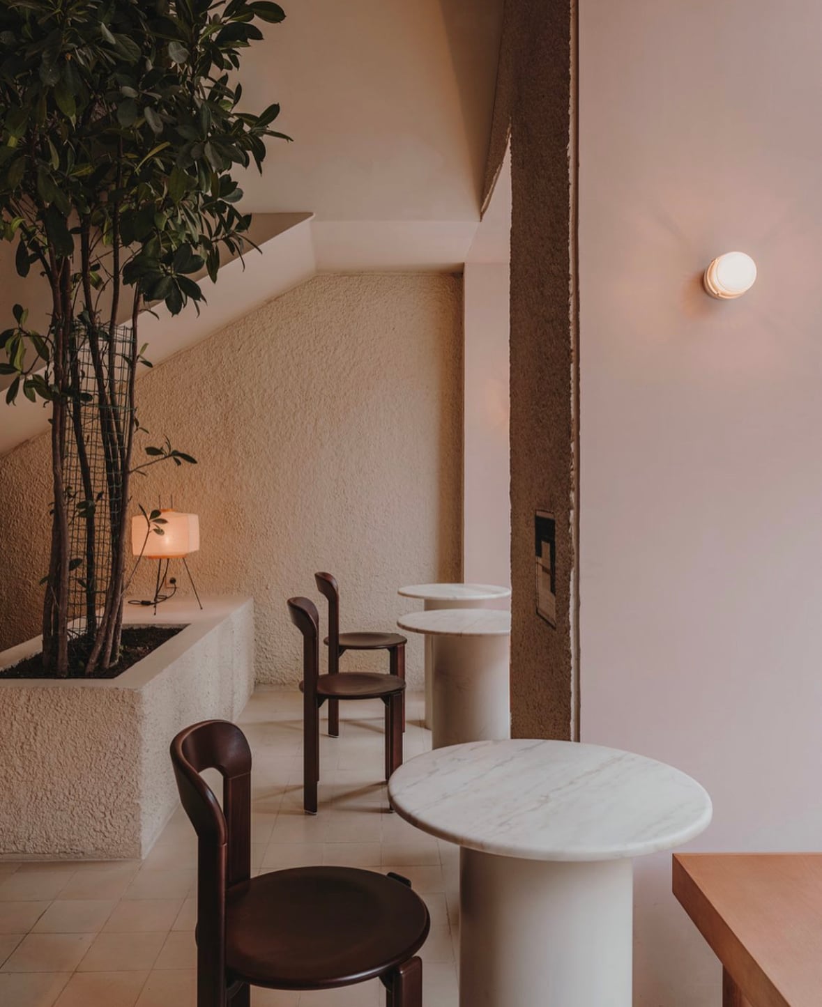 Llama Inn, Europe — Minimalist dining space with marble-topped cylindrical tables, dark wood chairs, textured cream walls, potted bamboo, and warm ambient lighting.