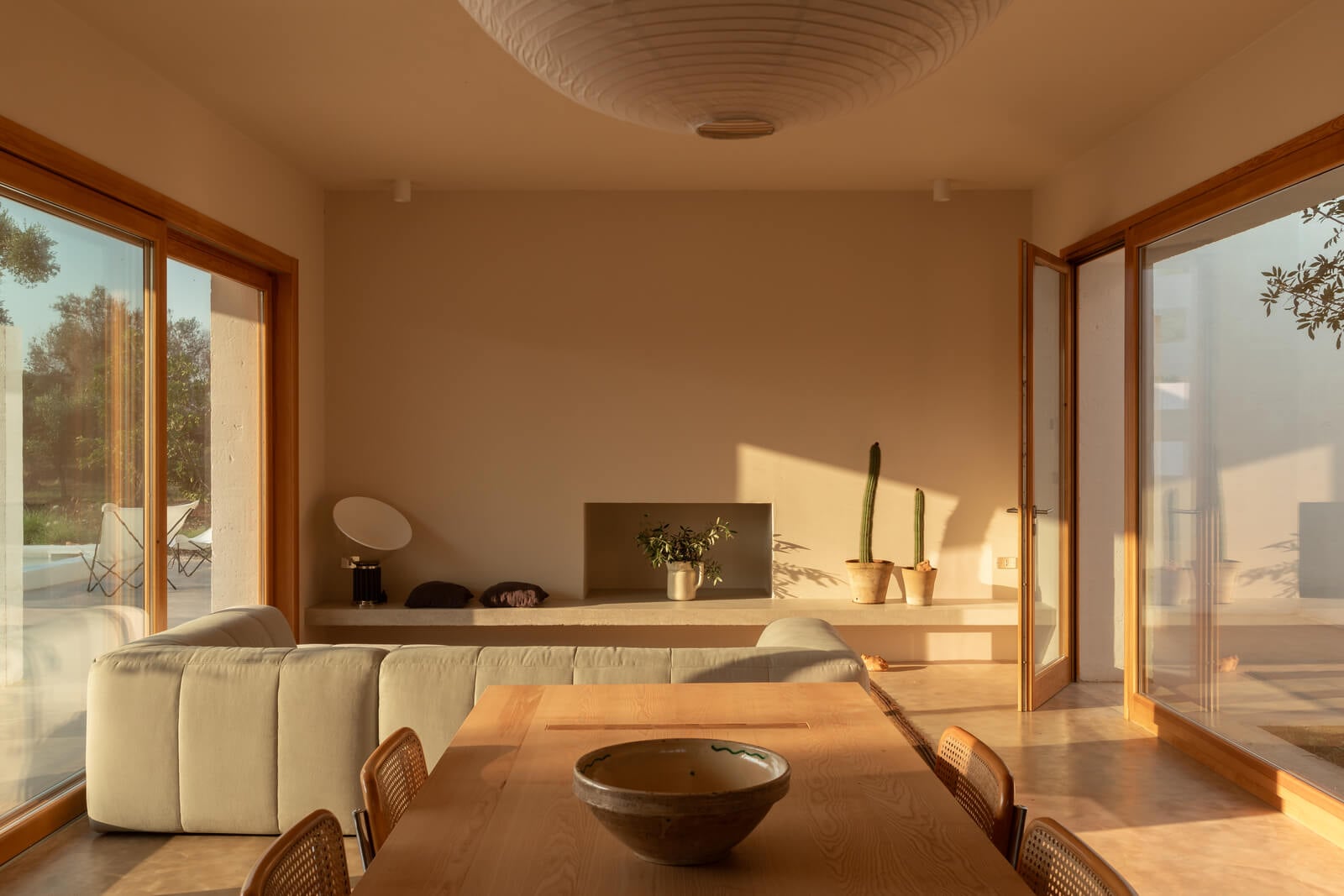 Casanguilla, Europe — Minimalist villa interior with curved cream sofa, wooden dining table, stone bowl, potted cacti, and warm sunlight through floor-to-ceiling windows.