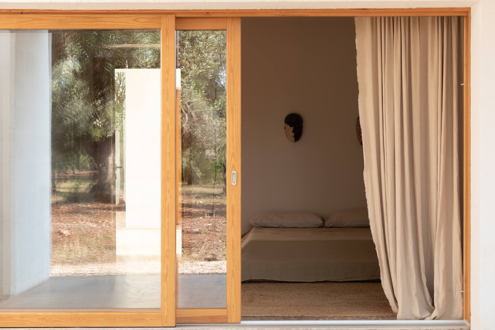 Casanguilla, Europe — Minimalist bedroom with wooden-framed glass doors opening to Mediterranean landscape, natural light, linen curtains, neutral palette.
