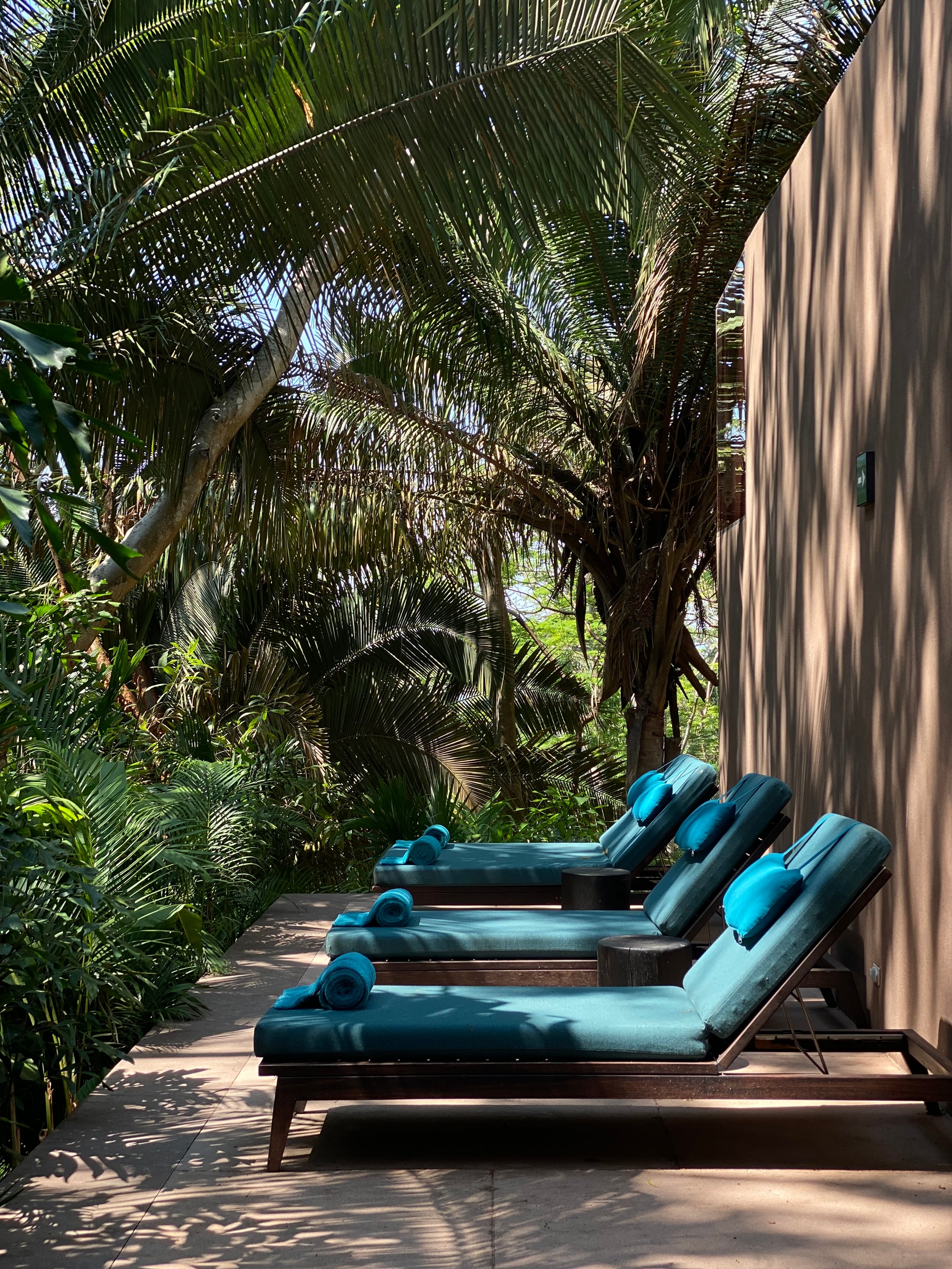 One&Only Mandarina, Mexico — Teak lounge chairs with blue cushions arranged on wooden deck beneath palm fronds, filtered sunlight casting shadows on corrugated metal wall.