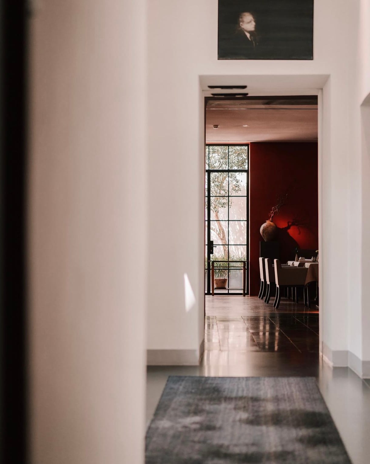 Purs, Europe — Minimalist hallway with polished concrete floor, white walls, gridded window framing foliage, and glimpse of red-walled dining room beyond.