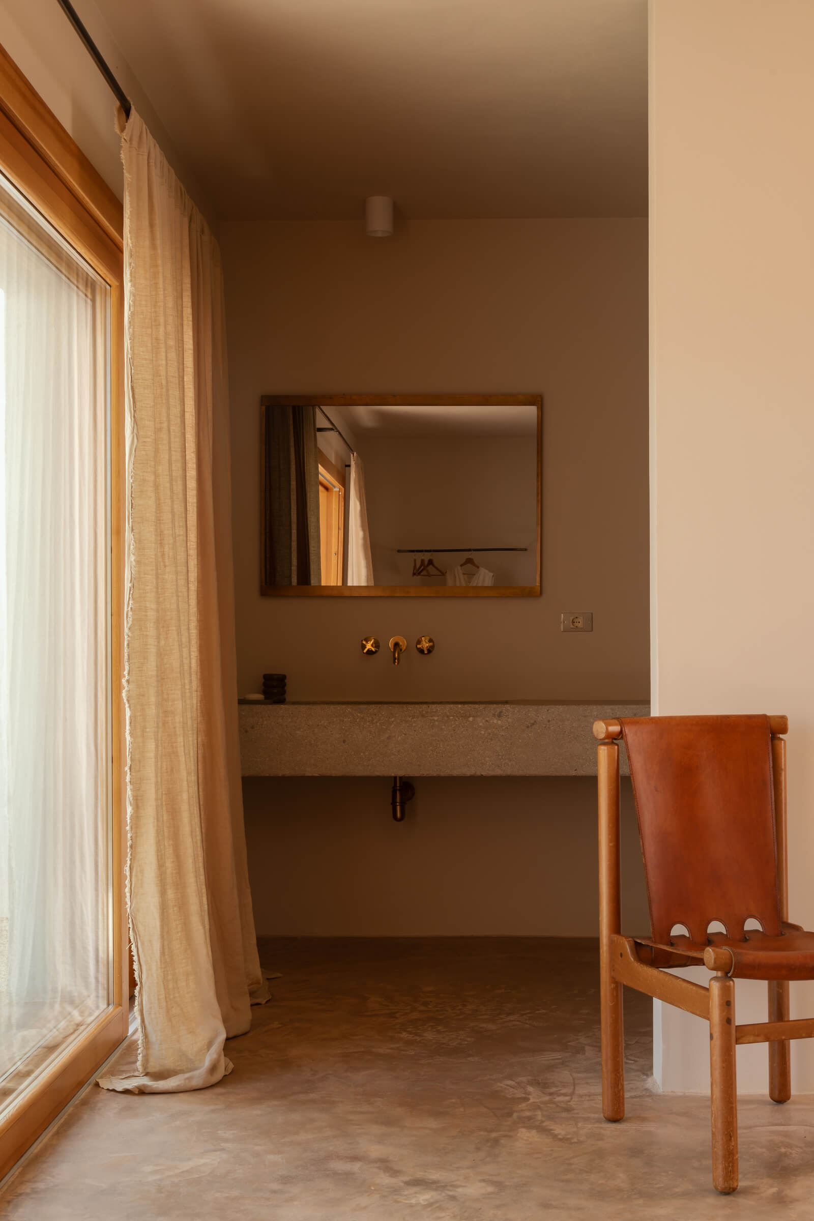 Casanguilla, Europe — Minimalist bathroom with terrazzo sink, wooden-framed mirror, brass fixtures, leather chair, and natural light from large window.