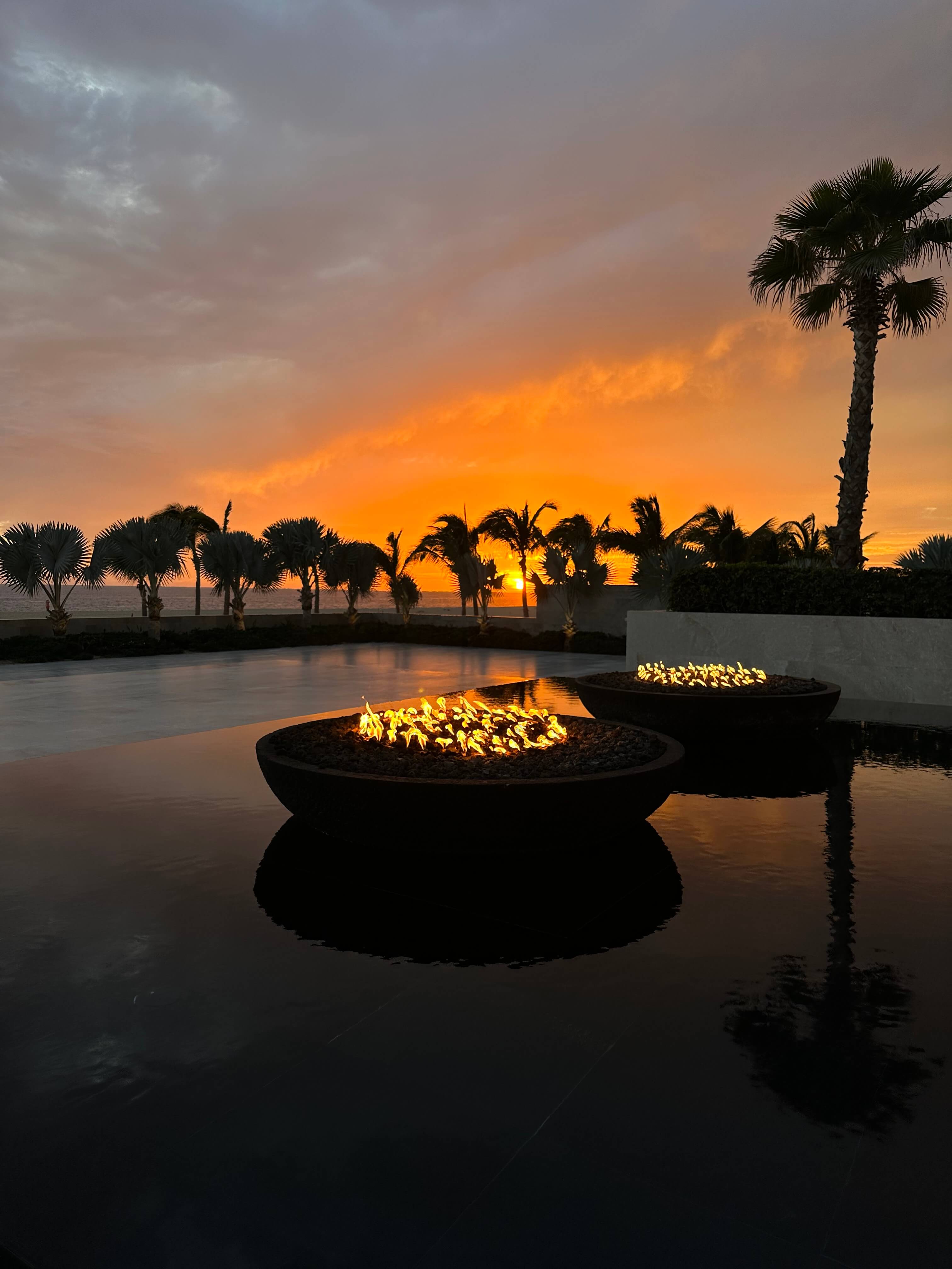 Nobu Los Cabos, Mexico — Illuminated fire bowls reflecting in still water at sunset, silhouetted palm trees against golden sky, modern luxury outdoor design.