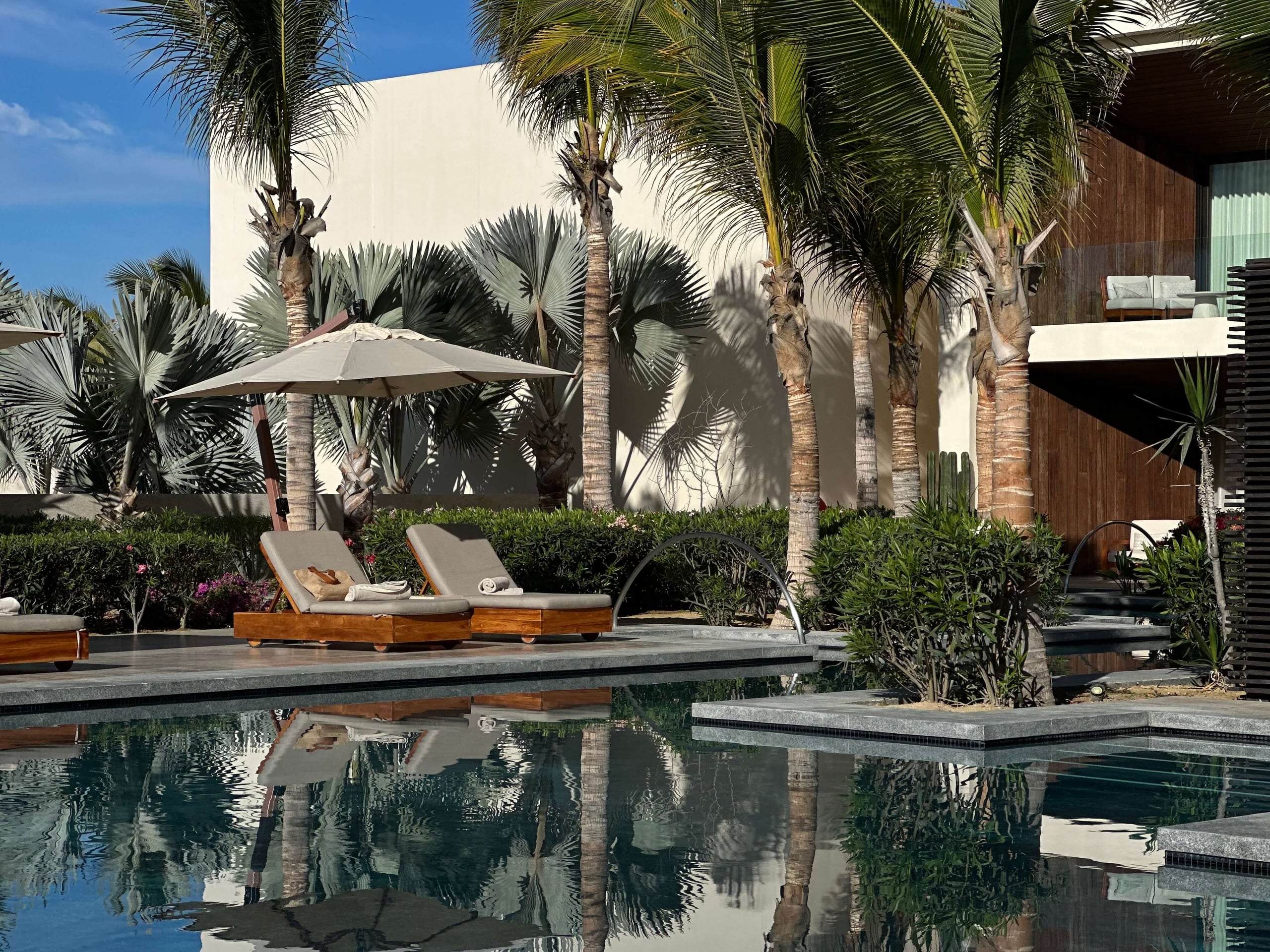Nobu Los Cabos, Mexico — Modernist poolside courtyard with teak loungers, gray umbrella, palm trees, reflecting pool, and minimalist white architecture under bright desert sun.