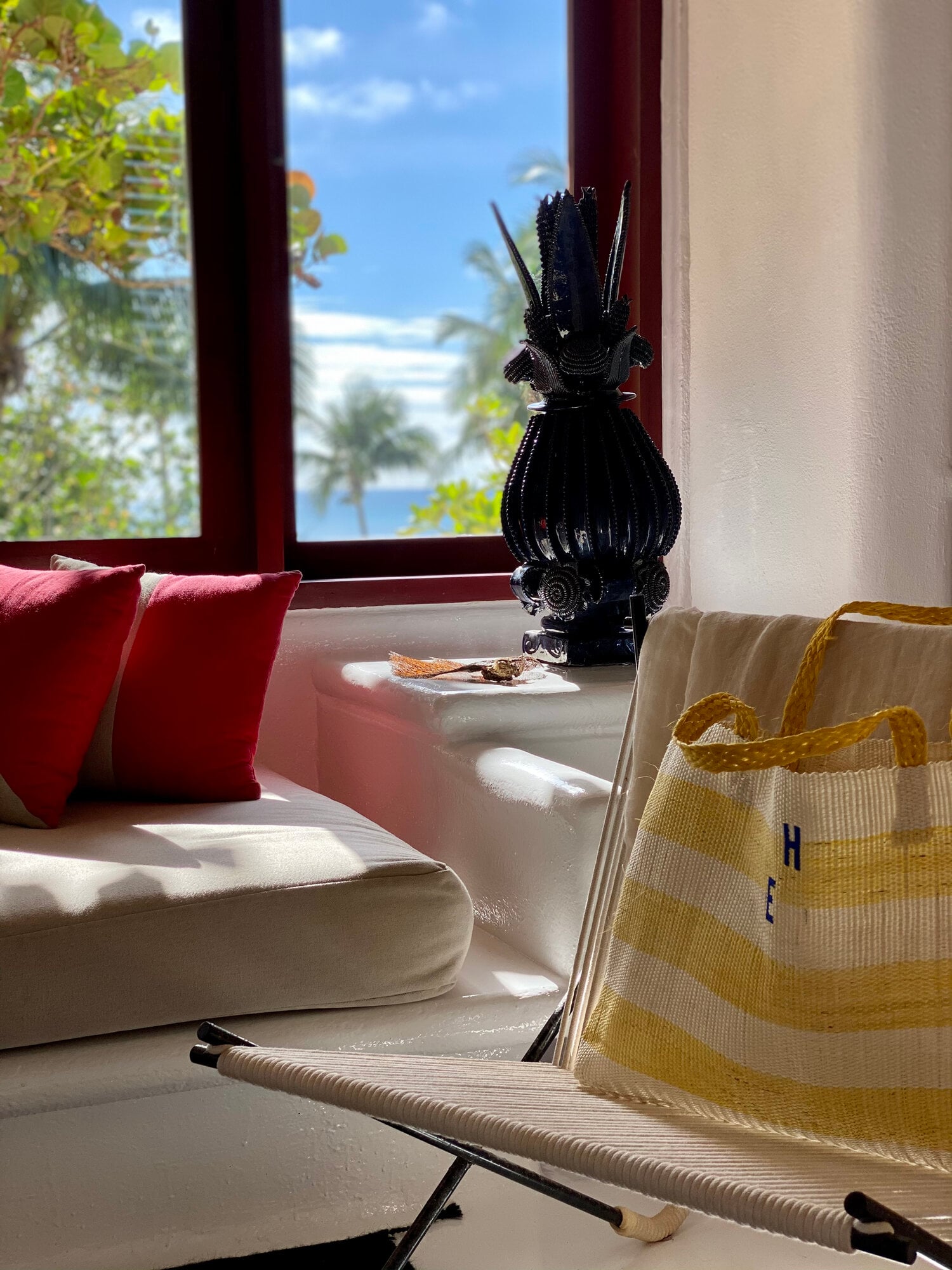 Sunlit windowsill at Hotel Esencia displaying red pillows, black sculptural vessel, and woven yellow tote against coastal backdrop.