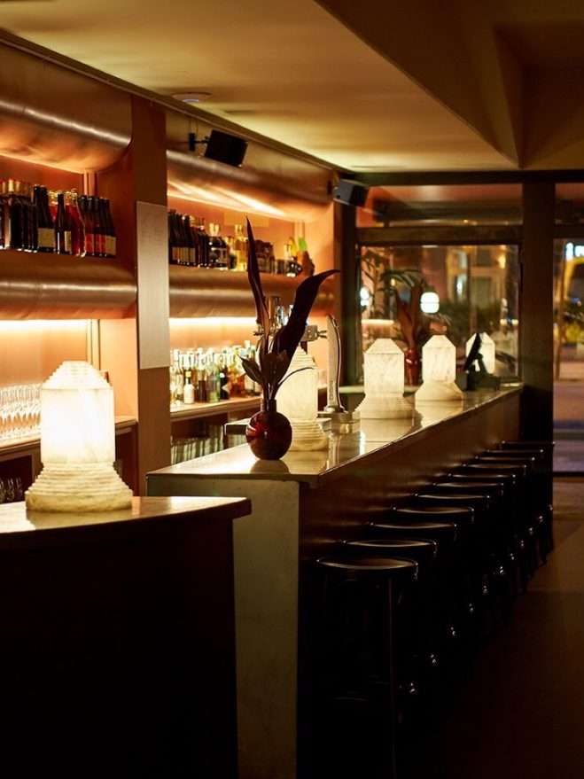 Casa Bonay, Europe — Illuminated bar interior with warm ambient lighting, dark bar stools, marble counter, backlit shelving, and botanical décor creating sophisticated hospitality design.