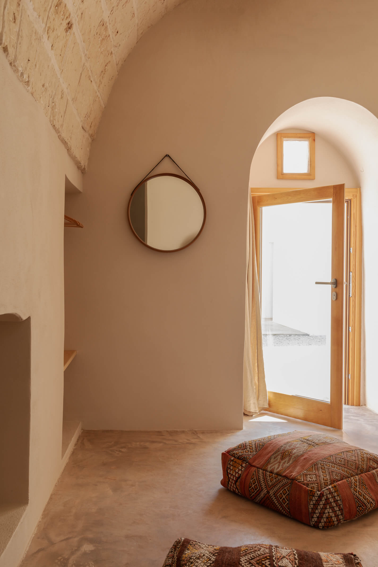 Casanguilla, Europe — Minimalist bedroom with terracotta walls, circular mirror, wooden door with arched threshold, and geometric patterned floor cushions.