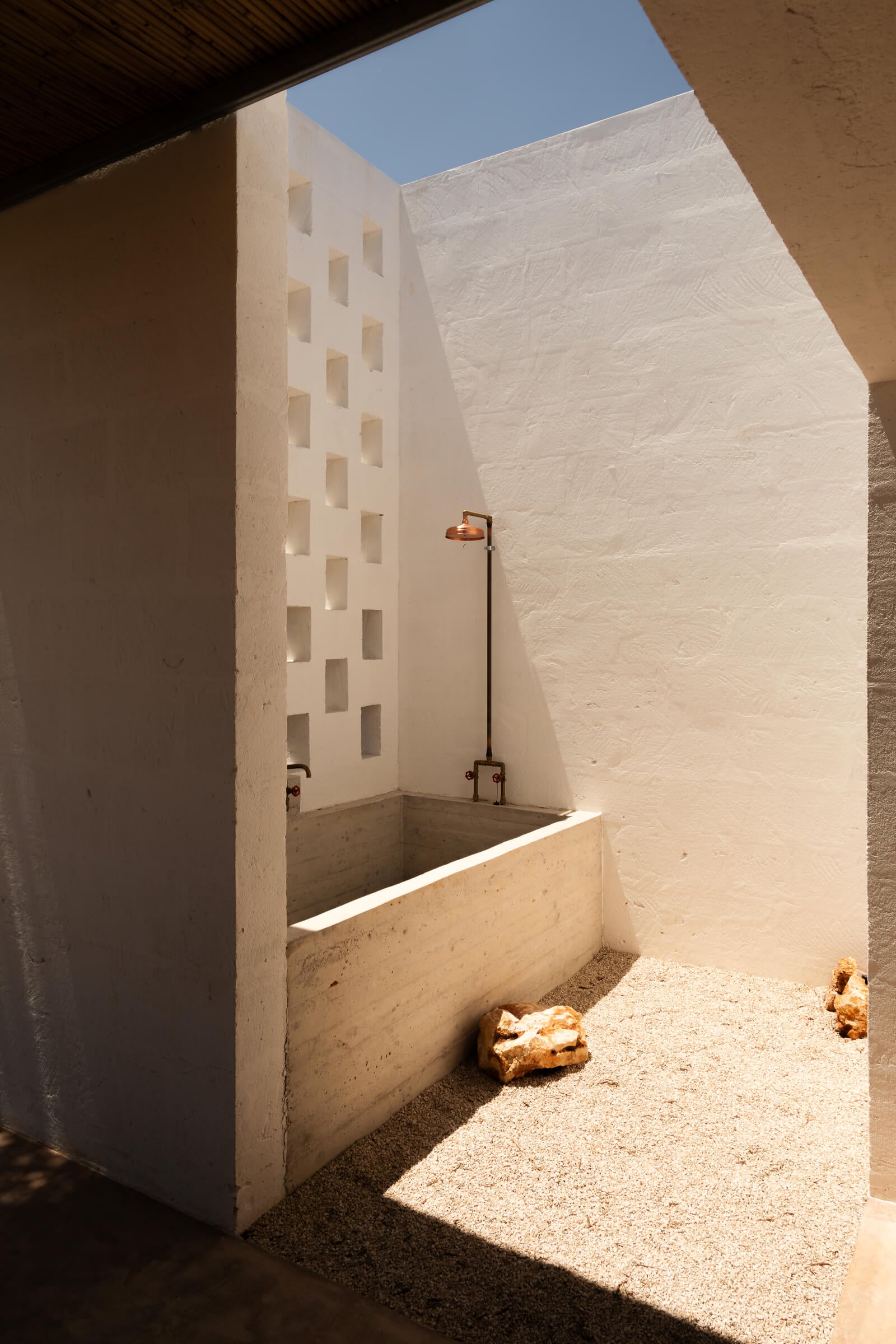 Casanguilla, Europe — Minimalist outdoor shower with whitewashed walls, ventilation blocks, concrete basin, and copper fixture under clear sky.
