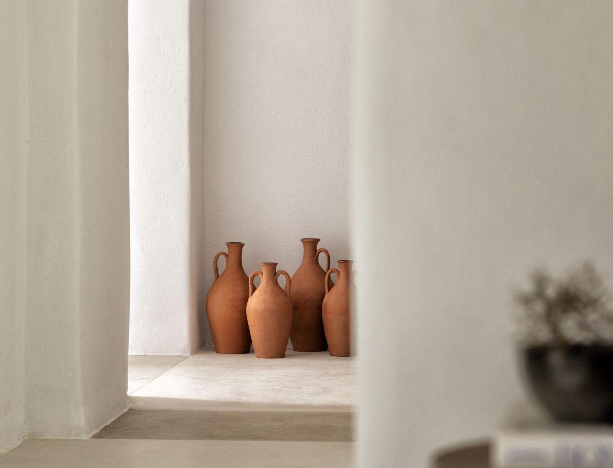 The Rooster, Europe — Terracotta amphoras arranged in minimalist white-walled niche with natural light, Mediterranean pottery styling.