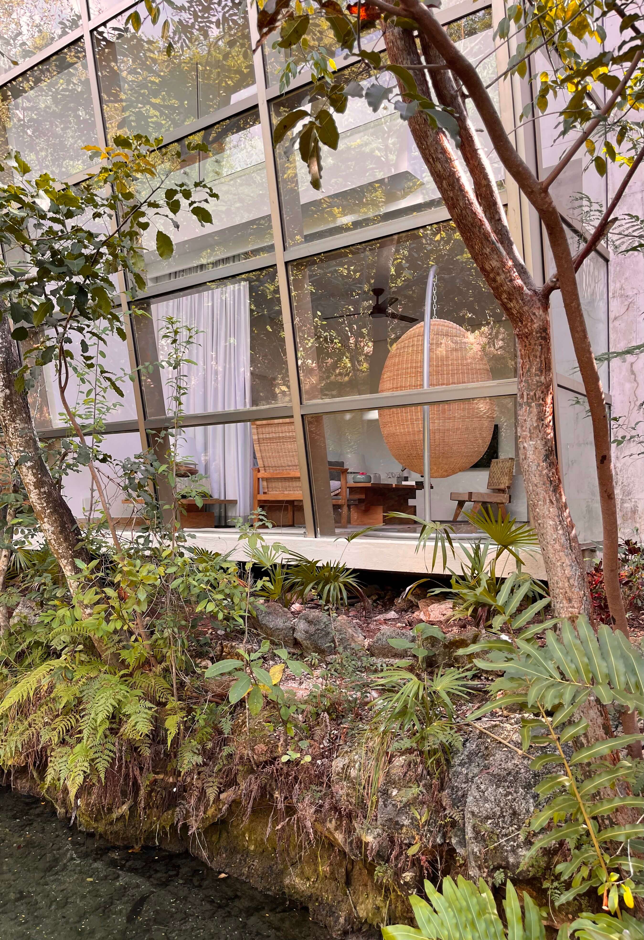 Rosewood Mayakoba, Mexico — Modernist glass pavilion with woven rattan furniture viewed through lush tropical garden with mature trees and native plants.