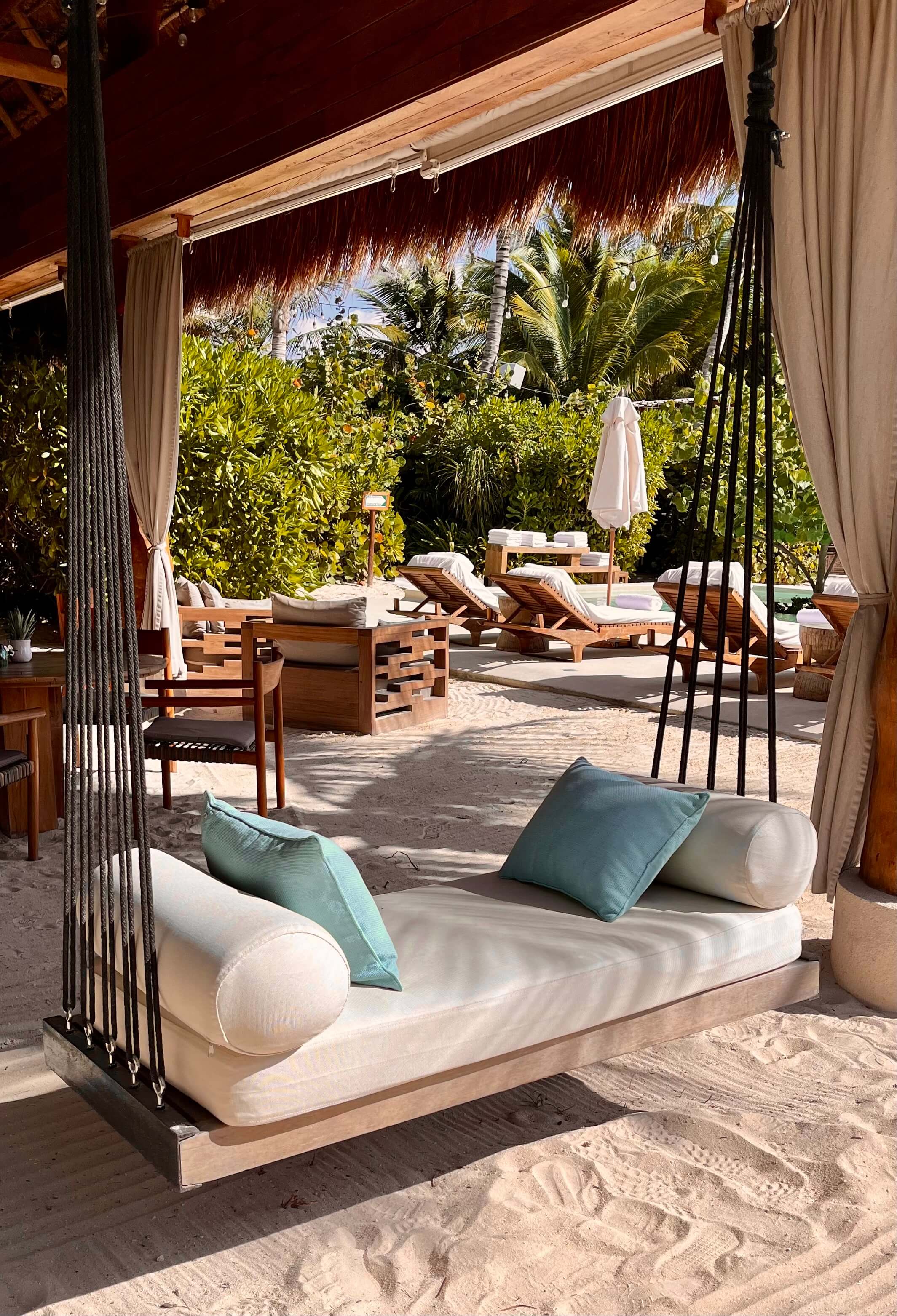 Rosewood Mayakoba, Mexico — Beachfront cabana with cream daybed, teak furniture, palapa roof, and lush tropical gardens overlooking Caribbean waters.