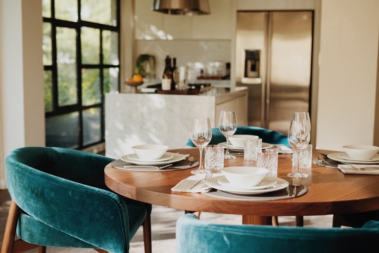 Campos Polanco, Mexico — Modern dining room with round wood table, teal velvet chairs, white place settings, crystal glasses, and open kitchen beyond tall windows.