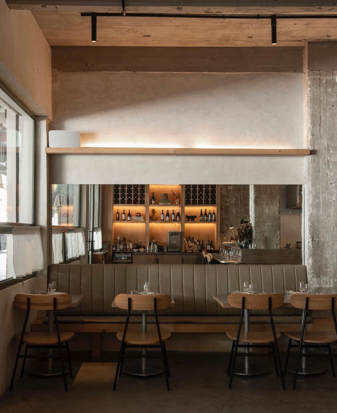 Martinez, Mexico — Modern restaurant interior with wood and black metal seating, concrete walls, warm-lit wine display, minimalist design, natural light from window.
