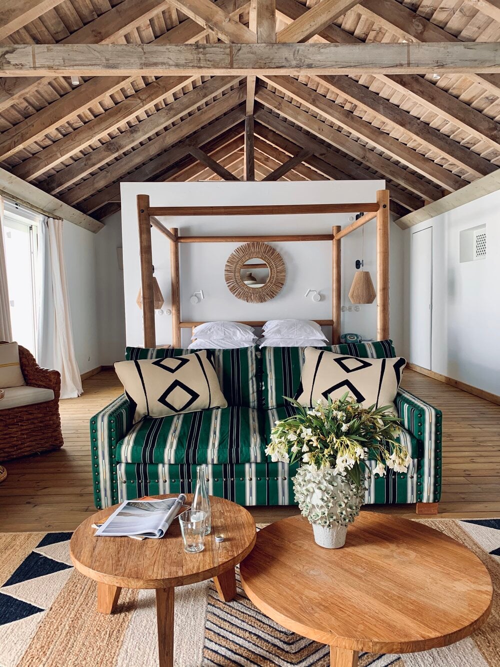 Quinta da Comporta, Europe — Bright bedroom with wooden four-poster bed, green striped sofa, exposed beam ceiling, round oak tables, and coastal minimalist design.
