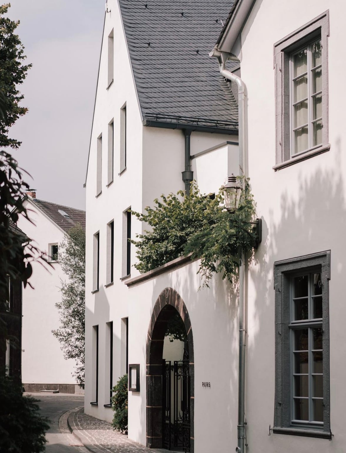 Purs, Europe — White-rendered archway with dark timber door, slate roof, ivy-covered courtyard, modernist minimalist architecture with natural light and stone paving.