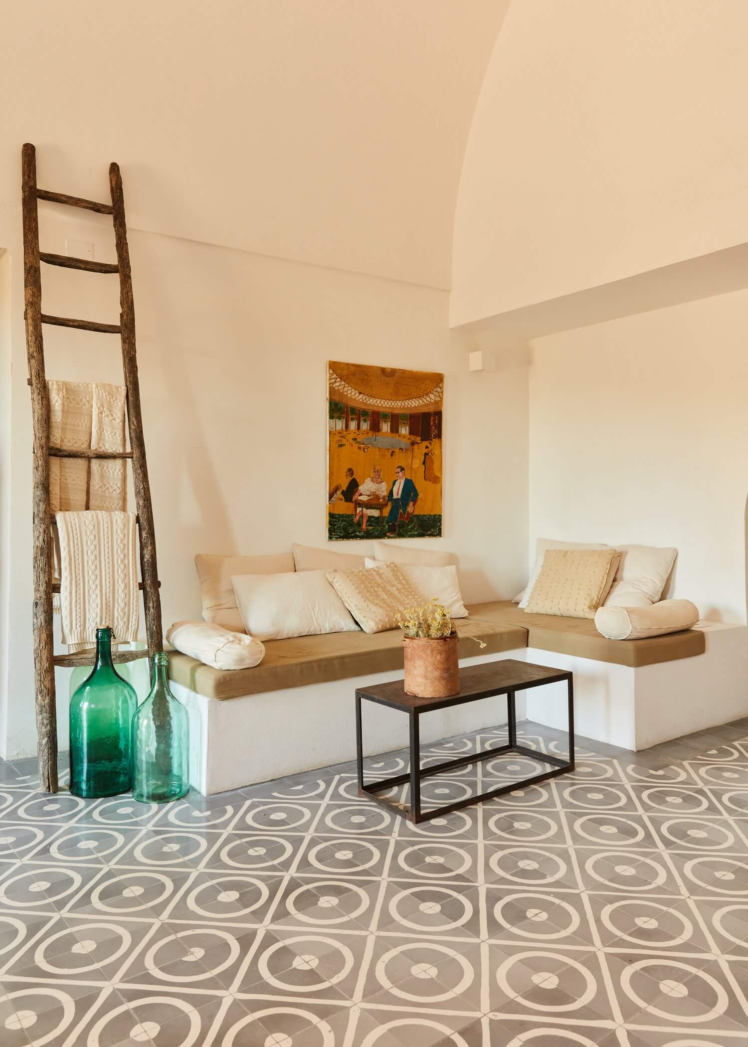 Parco Dei Sesi, Europe — Minimalist lounge with cream sectional sofa, geometric patterned floor, wooden ladder, vintage green bottles, and ochre artwork beneath vaulted ceiling.