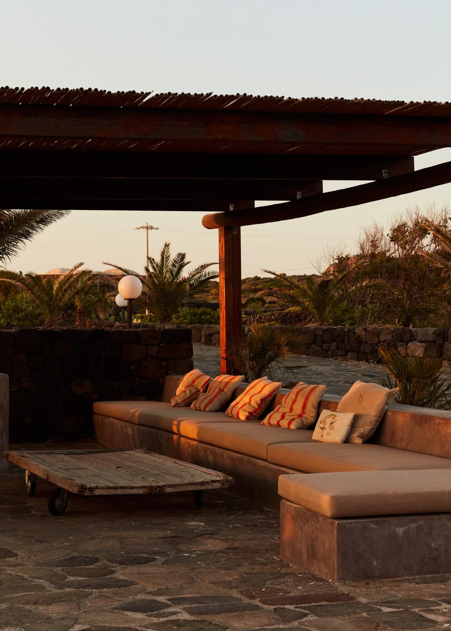 Parco Dei Sesi, Europe — Wooden pergola terrace at sunset with earth-tone seating, striped pillows, stone walls, and desert landscape views.