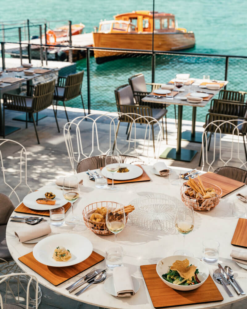 Il Sereno Al Lago, Europe — Waterfront dining terrace with round white table, wooden accents, woven chairs, fresh pasta dishes, turquoise water, wooden boat, Mediterranean coastal setting.
