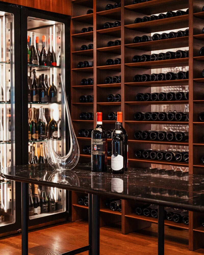 Il Sereno Wine Cellar, Europe — Illuminated wine storage with wooden shelves, black marble counter, glass-front refrigerator, and two featured bottles under warm ambient lighting.