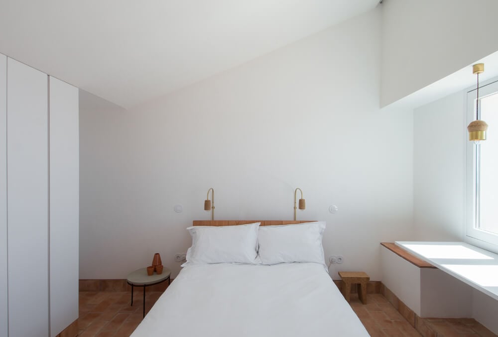 Casa Modesta, Europe — Minimalist bedroom with white bedding, wooden headboard, brass pendant lights, terracotta flooring, and natural window light.
