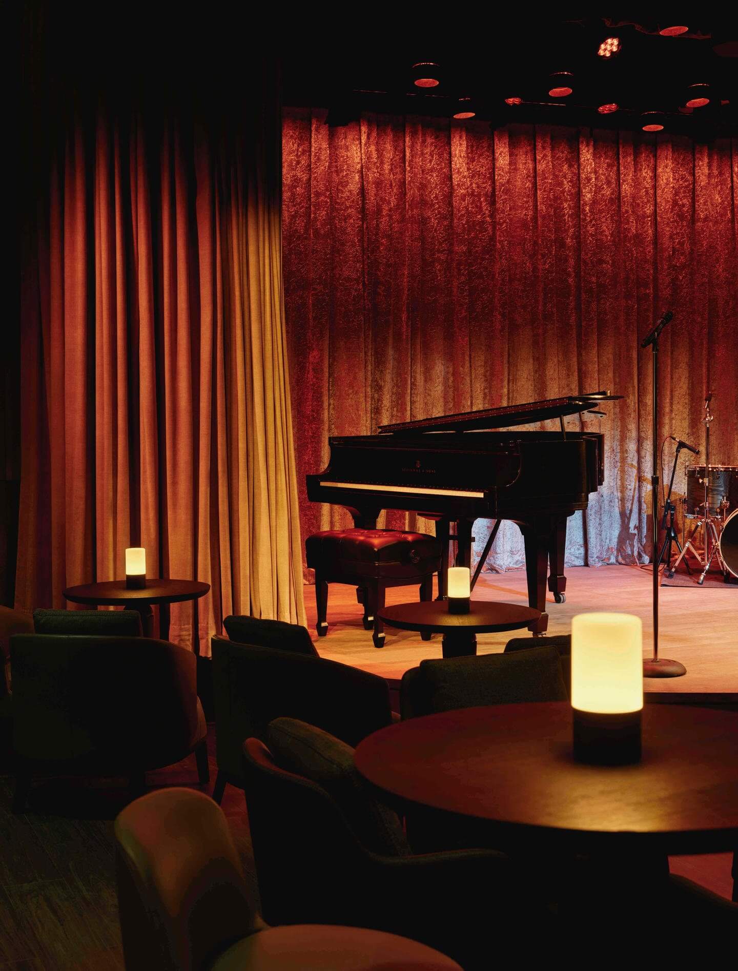 The Jazz Club, North America — Grand piano on warm-lit stage with red velvet curtains, intimate lounge seating, and glowing cylindrical accent lights.