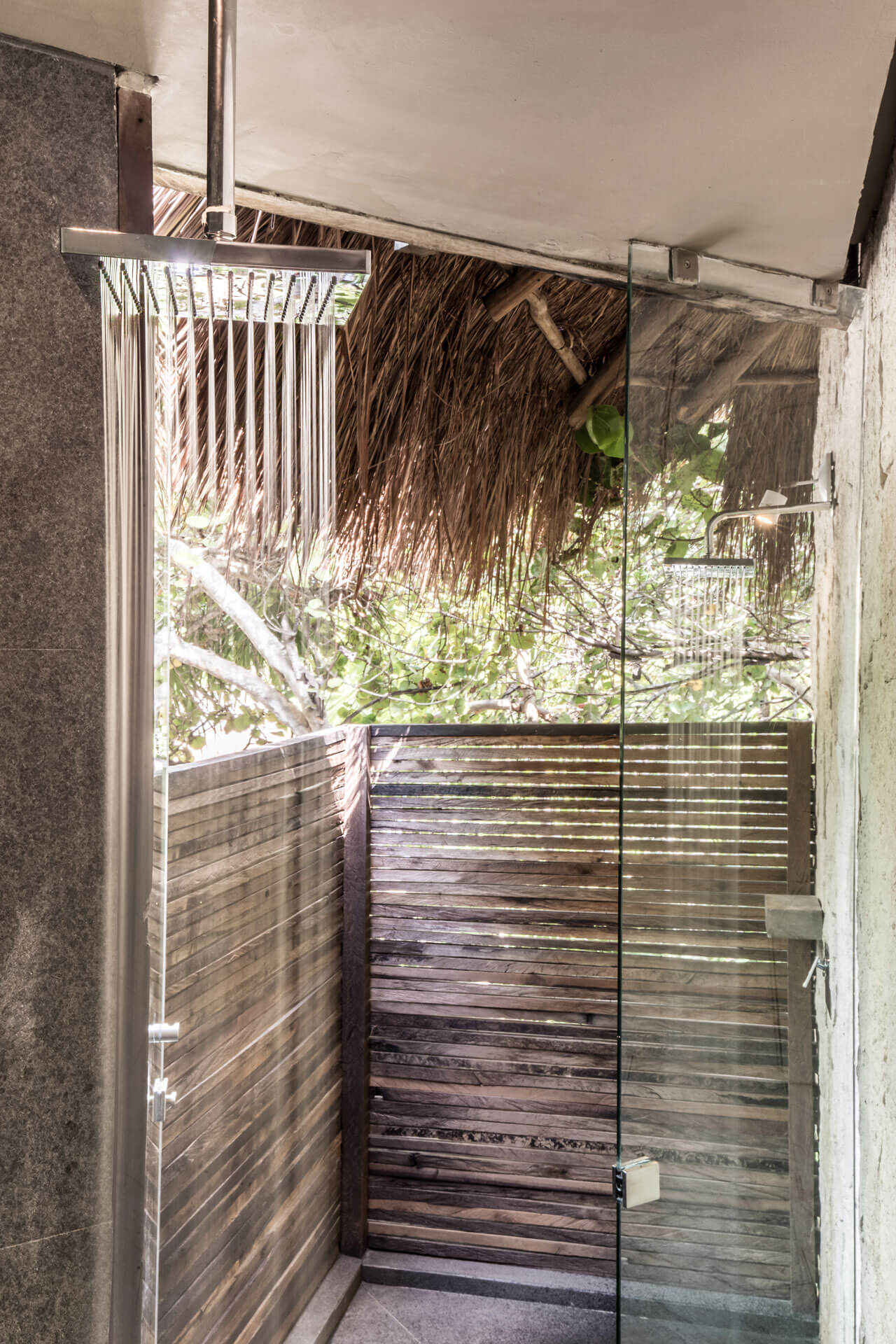 Be Tulum, Mexico — Outdoor shower with thatched roof, glass enclosure, wooden deck, and lush tropical vegetation viewed through open door.
