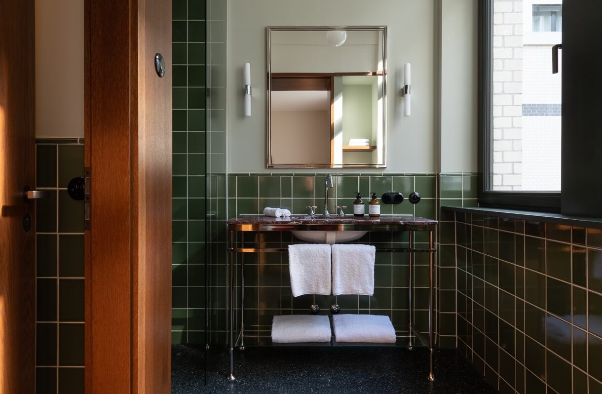 Château Royal, Europe — Art Deco bathroom with forest-green tile, brass vanity, marble countertop, framed mirror, and soft ambient lighting through window.
