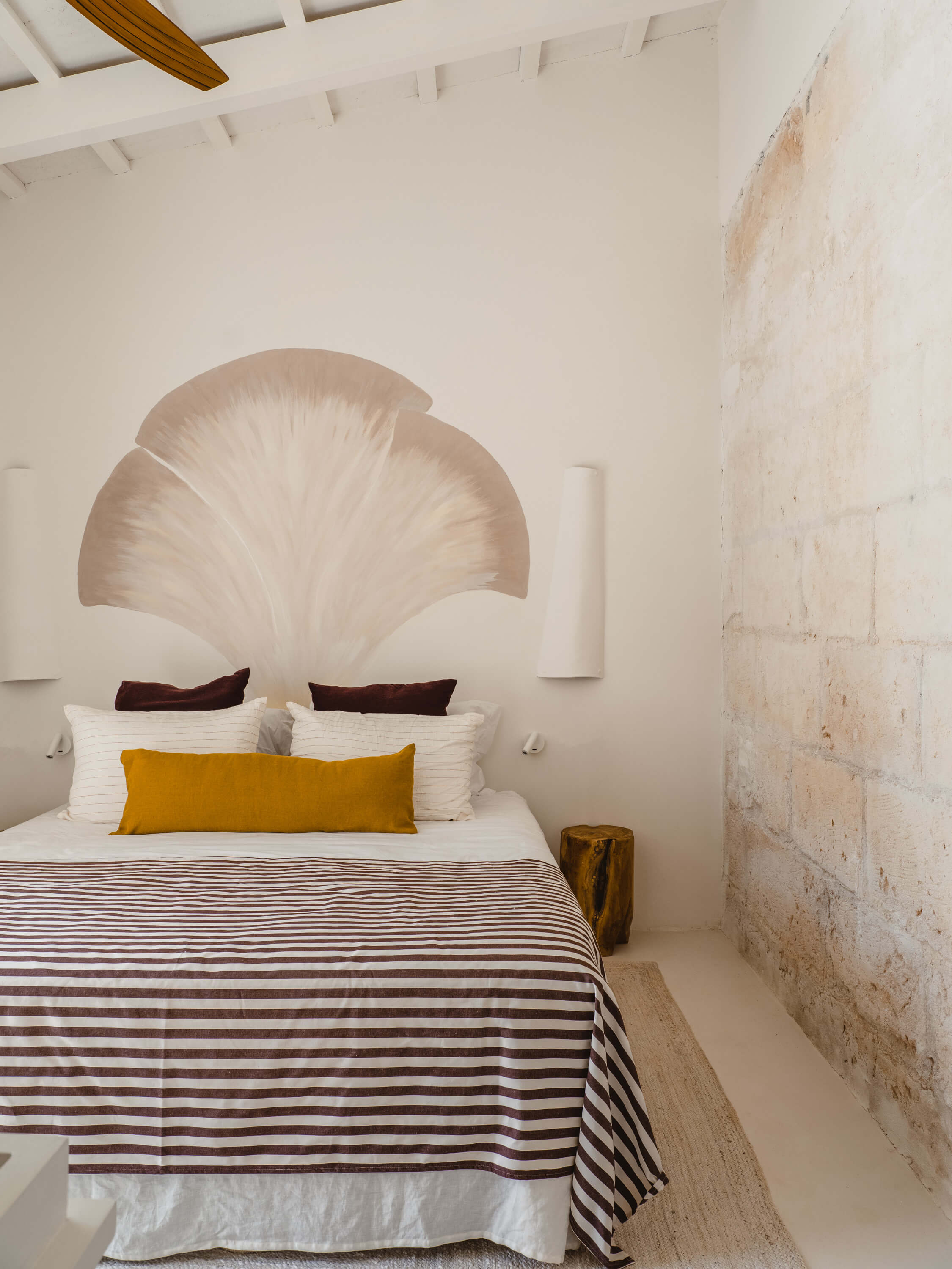Morvedra Nou, Balearic Islands — Minimalist bedroom with curved shell headboard, striped bedding, golden pillow, and whitewashed stone walls.