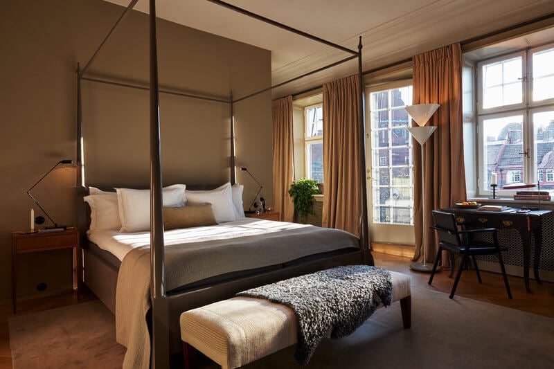 Ett Hem, Europe — Canopy bed with black frame in warm-toned bedroom, floor-to-ceiling windows overlooking cityscape, ochre curtains, wooden desk, contemporary luxury hotel interior.