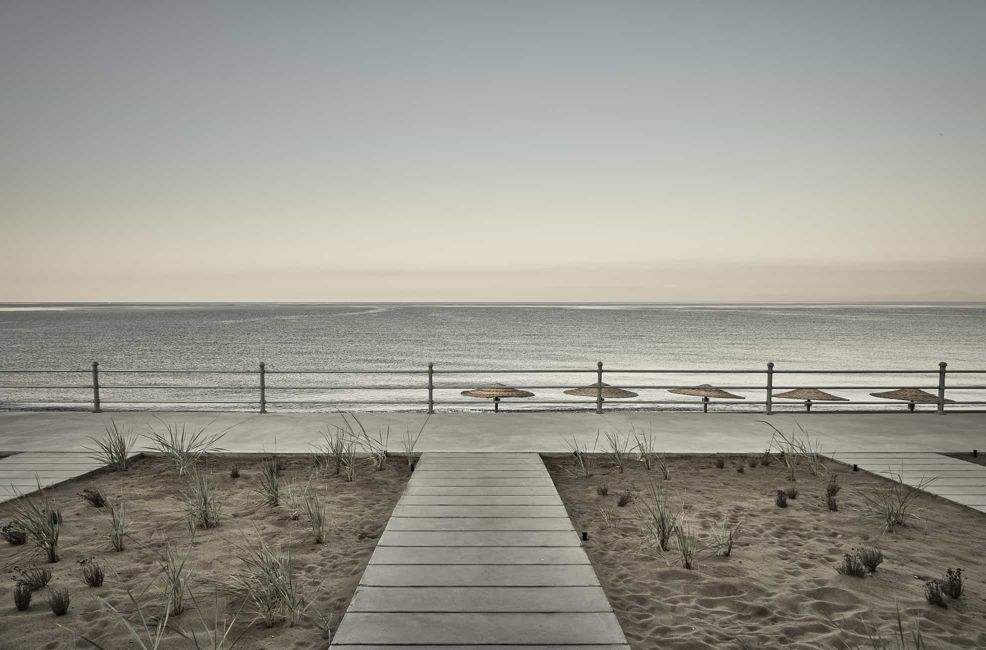 Dexamenes Seaside Hotel, Europe — Minimalist boardwalk with umbrellas overlooking calm sea, dune grasses, and metal railings at dusk.
