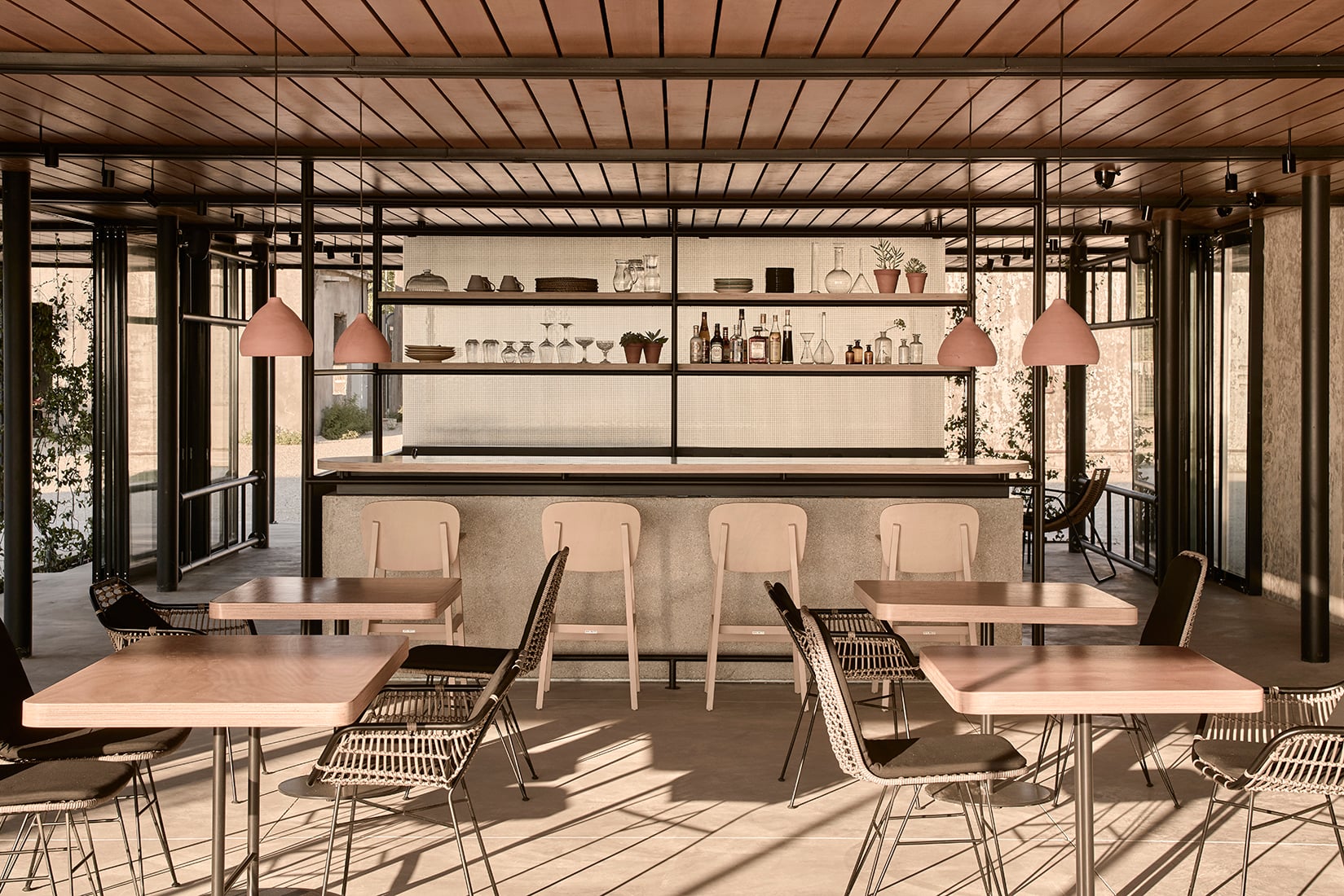 The Beachfront Bar — Beachfront Bar, Europe — Modern pavilion with timber slatted ceiling, concrete bar, woven rattan chairs, pink pendant lights, and floor-to-ceiling glass overlooking garden terrace.