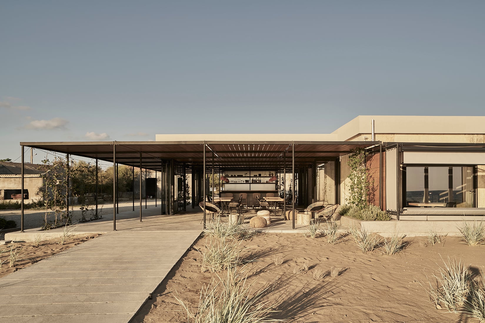 Dexamenes Seaside Hotel, Europe — Modern minimalist beachfront pavilion with steel pergolas, sand landscaping, and floor-to-ceiling glazing overlooking Mediterranean coastline.