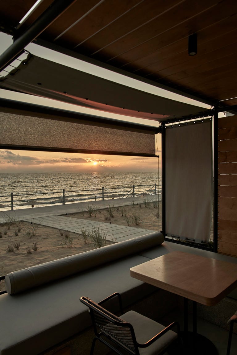 Dexamenes Seaside Hotel, Europe — Minimalist workspace with rolling shade overlooking sunset beach vista, wooden ceiling, contemporary furniture, golden hour light.