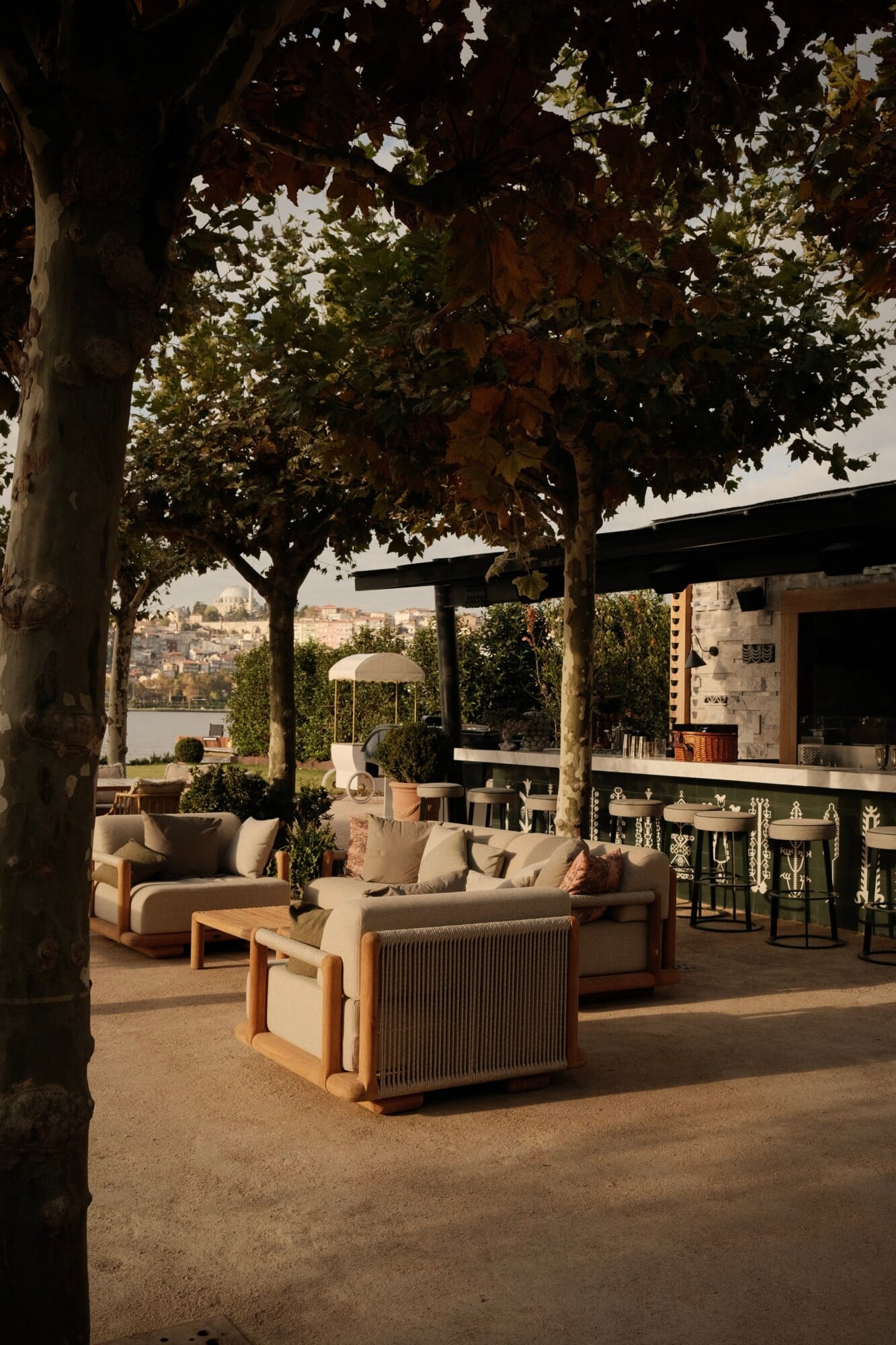 Hasbahçe, Istanbul, Turkey — Shaded waterfront terrace with cream upholstered seating, wooden pergola, mature plane trees, and panoramic Golden Horn views.