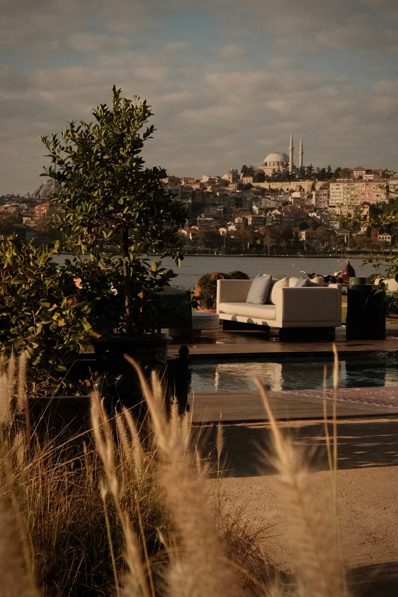 Aliée Istanbul, Istanbul, Turkey — Waterfront lounge with cream seating, olive plants, and Golden Horn views toward mosque and minaret at golden hour.