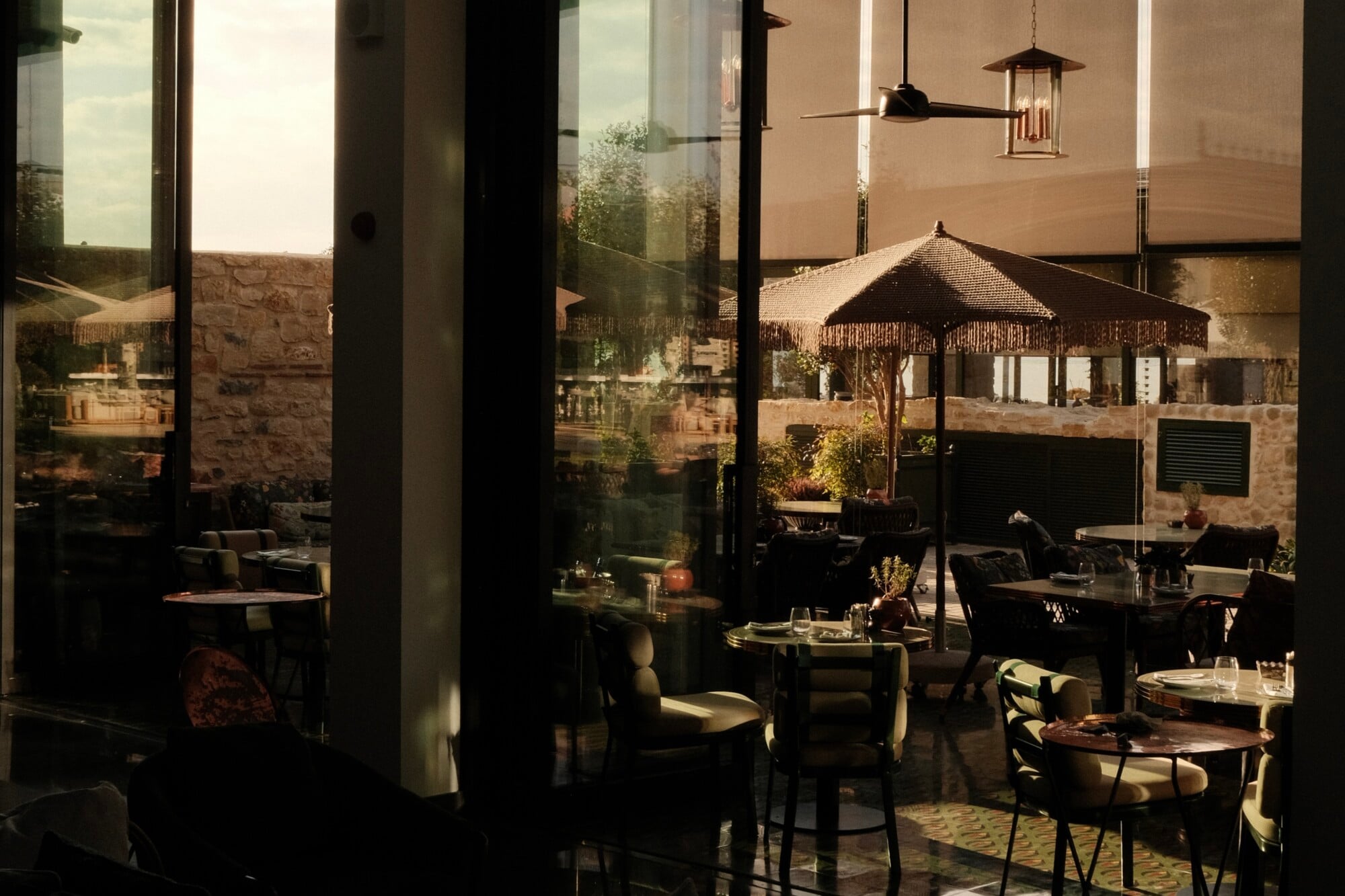Aliée Istanbul, Istanbul, Turkey — Sunlit courtyard dining room with stone walls, dark columns, woven umbrella, and modern pendant lighting overlooking landscaped gardens.