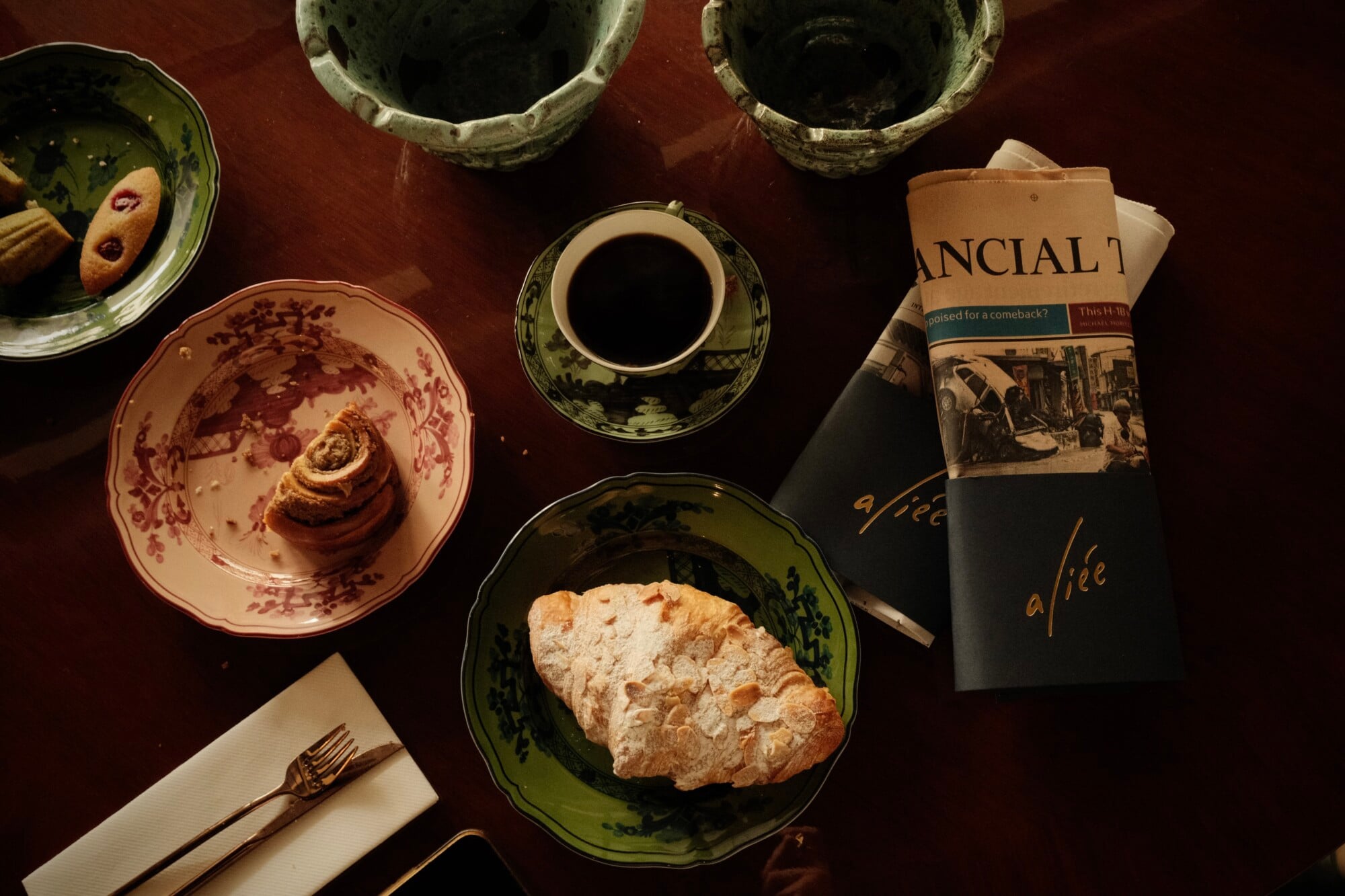 Aliée Istanbul, Turkey — Overhead table setting with pastries, vintage porcelain, black coffee, and newspaper on deep burgundy surface, intimate morning scene.