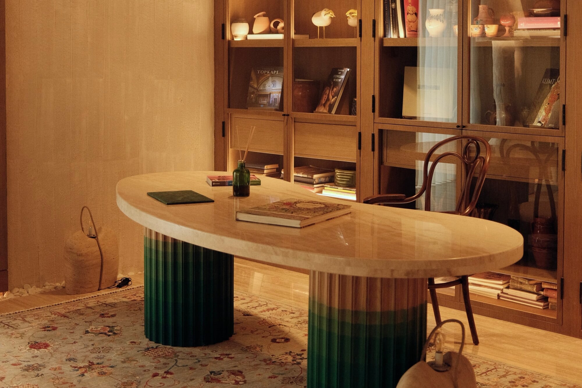 Aliée Istanbul, Turkey — Oval wooden desk with green fluted base in warm-lit study, brass chairs, built-in cabinetry with books and ceramics behind.