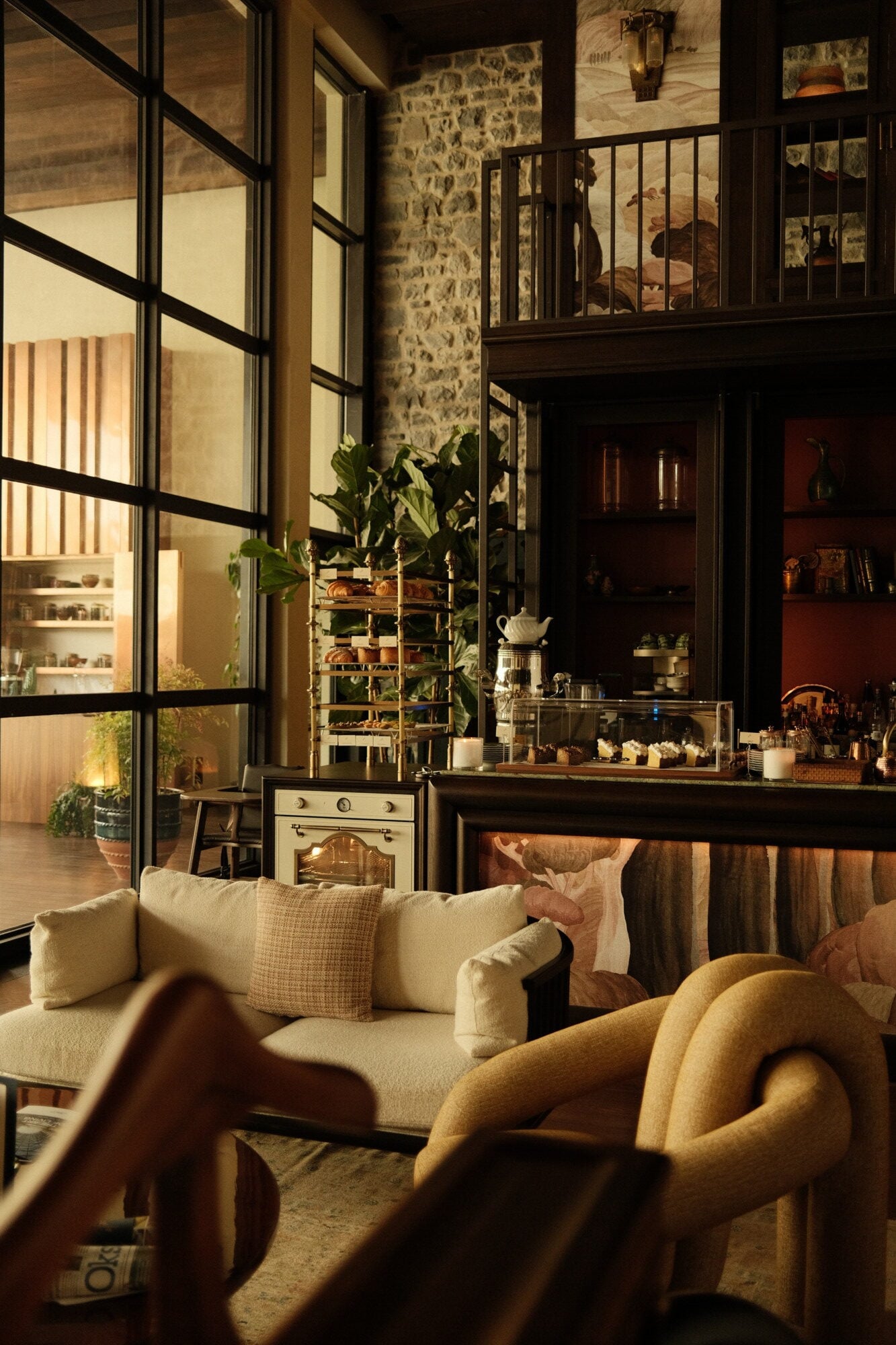 Aliée Istanbul, Istanbul, Turkey — Cream sofa with golden armchairs facing floor-to-ceiling windows, stone walls, and bar shelving in warm evening light.