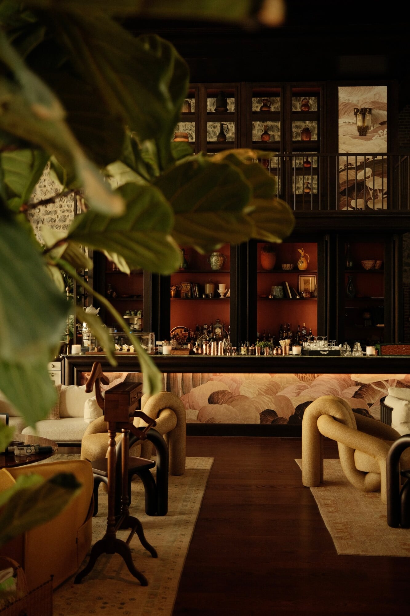 Aliée Istanbul, Turkey — Warm interior with curved sculptural furniture, terracotta shelving, potted plant foreground, and curated photographs on upper gallery wall.