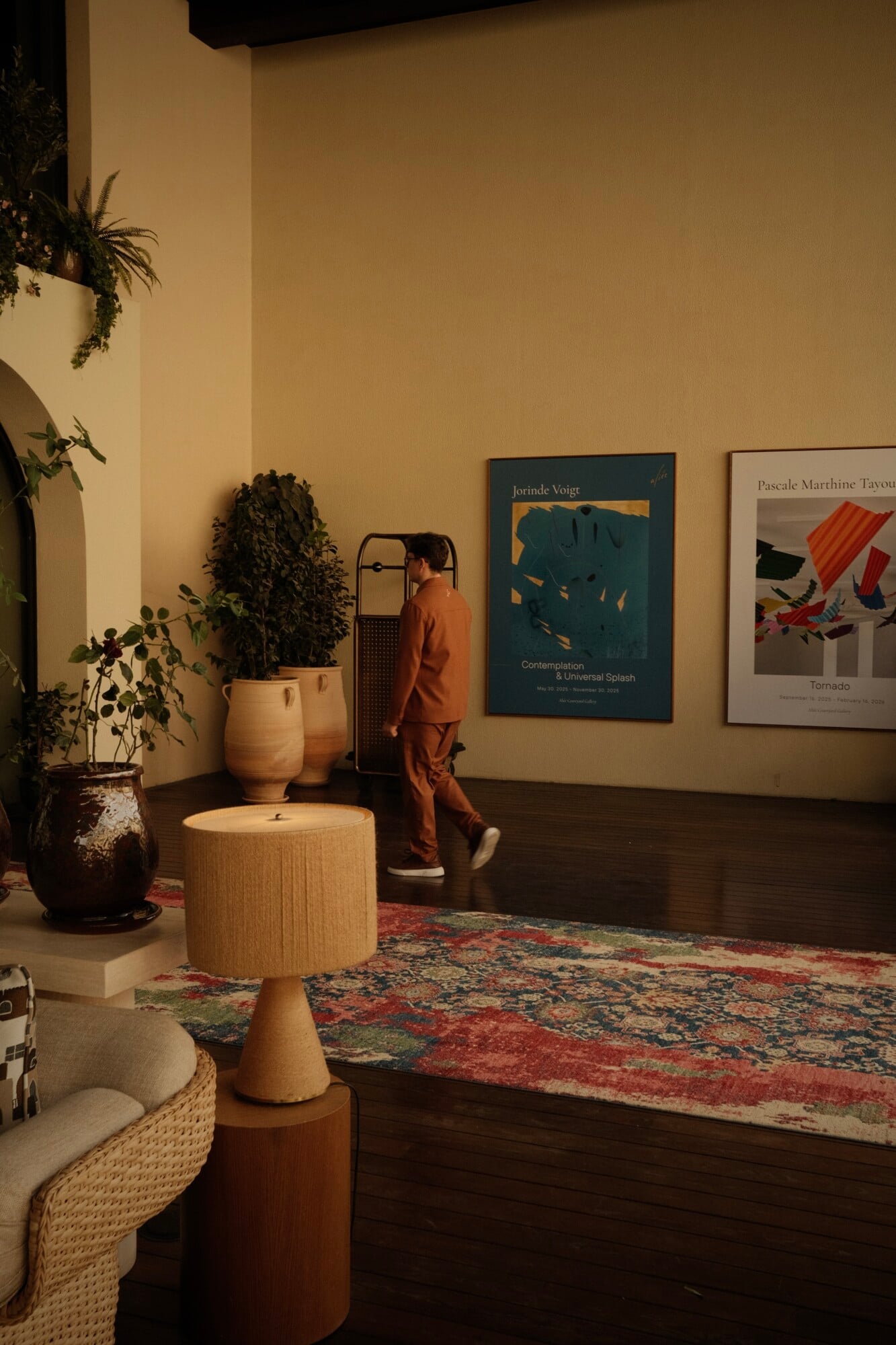 A contemplative figure walks through a warm, gallery-like interior at Aliée Istanbul, surrounded by natural wood furniture, potted plants, and contemporary art on pale walls.