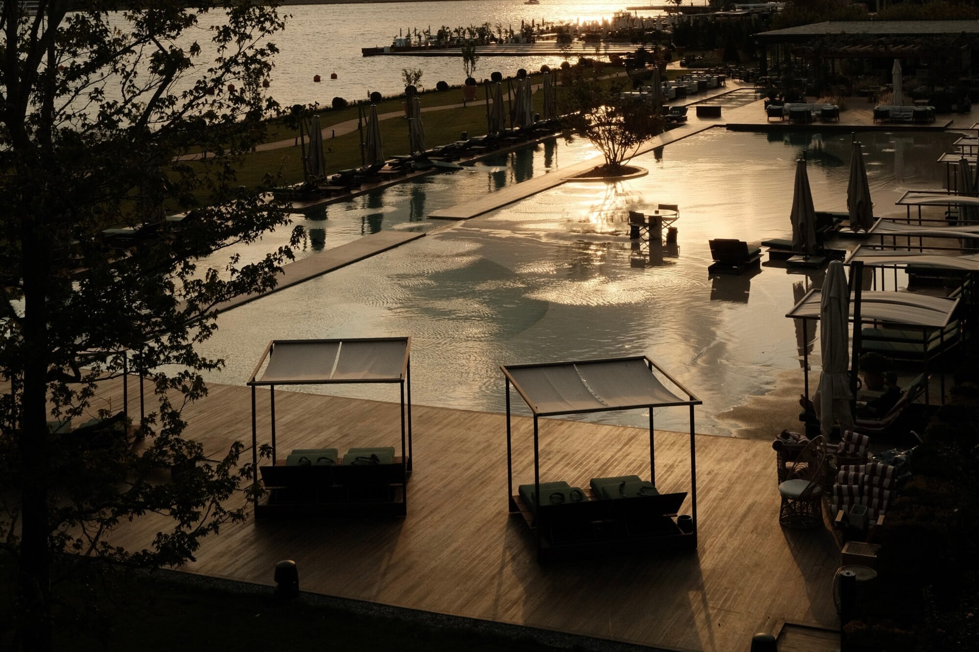 Aliée Istanbul, Turkey — Waterfront terrace at golden hour with canopied lounge beds, wooden decking, and calm harbor views reflecting soft light.