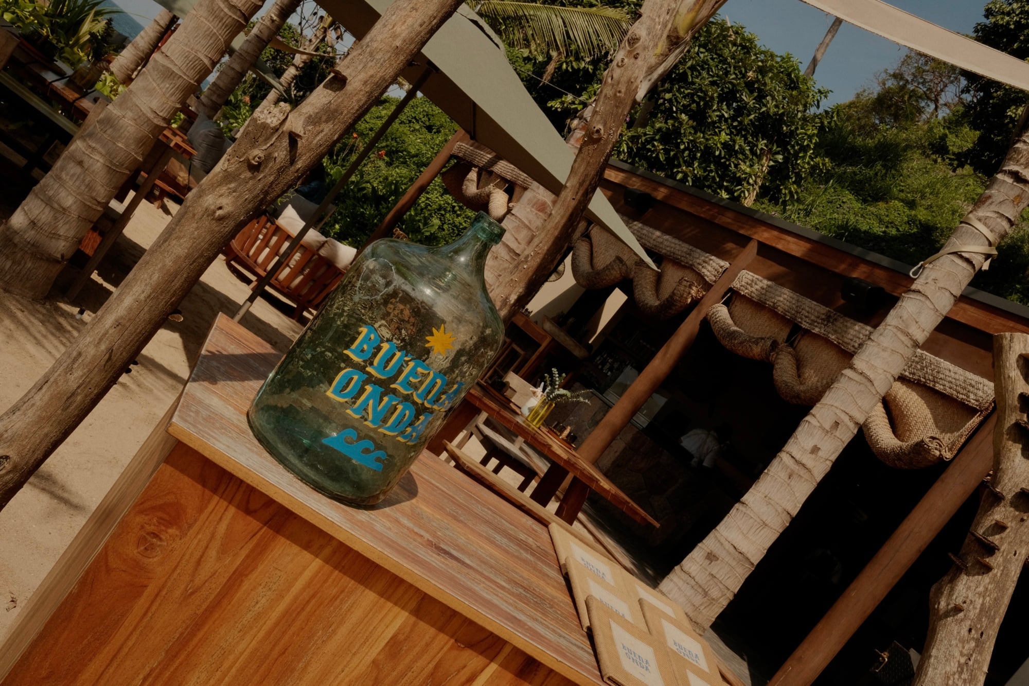 Buena Onda, Mexico — Rustic wooden bar with vintage glass bottle and palm wood pergola structure overlooking lush garden seating area.