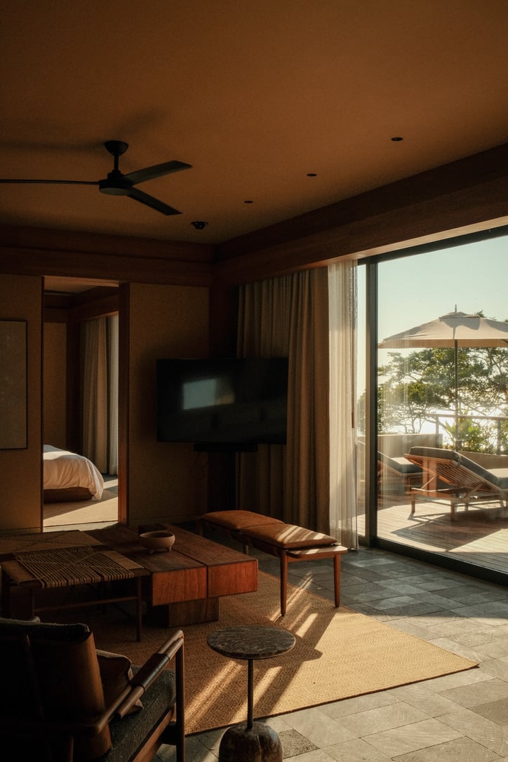 Rosewood Mandarina, Mexico — Warm-toned resort suite with mid-century wooden furniture, floor-to-ceiling windows overlooking ocean terrace, natural light casting shadows across stone flooring.