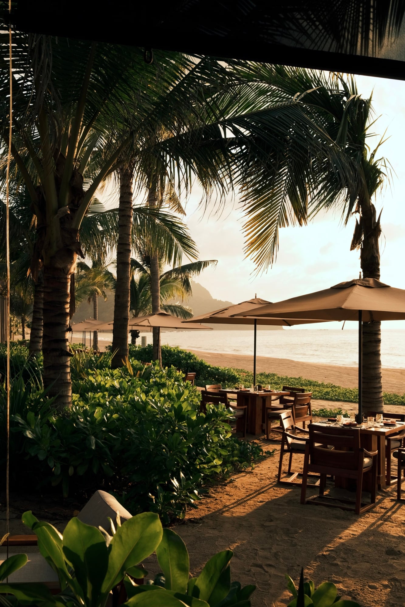 Rosewood Mandarina, Mexico — Beach pavilion with timber dining tables, canvas umbrellas, and palm fronds backlit against ocean and coastal mountains at golden hour.